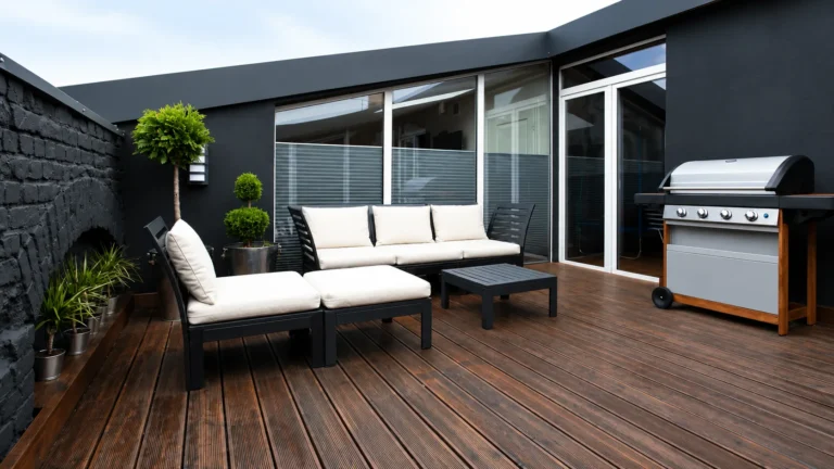 Modern outdoor patio with wood decking, lounge furniture, large windows, potted plants, and barbecue grill.