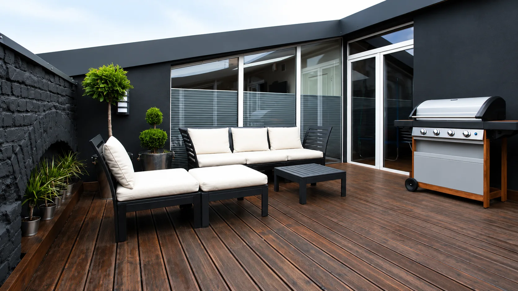 Modern outdoor patio with wood decking, lounge furniture, large windows, potted plants, and barbecue grill.